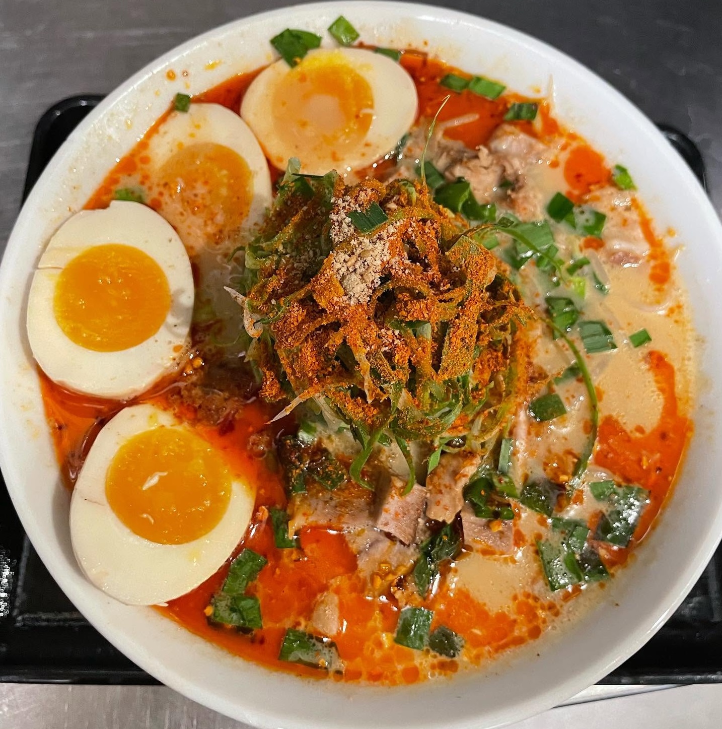 A bowl of spicy ramen noodles Costa Mesa topped with four halved soft-boiled eggs, sliced green onions, tender pork pieces, chili powder, and a creamy, orange-tinged broth with oil droplets.