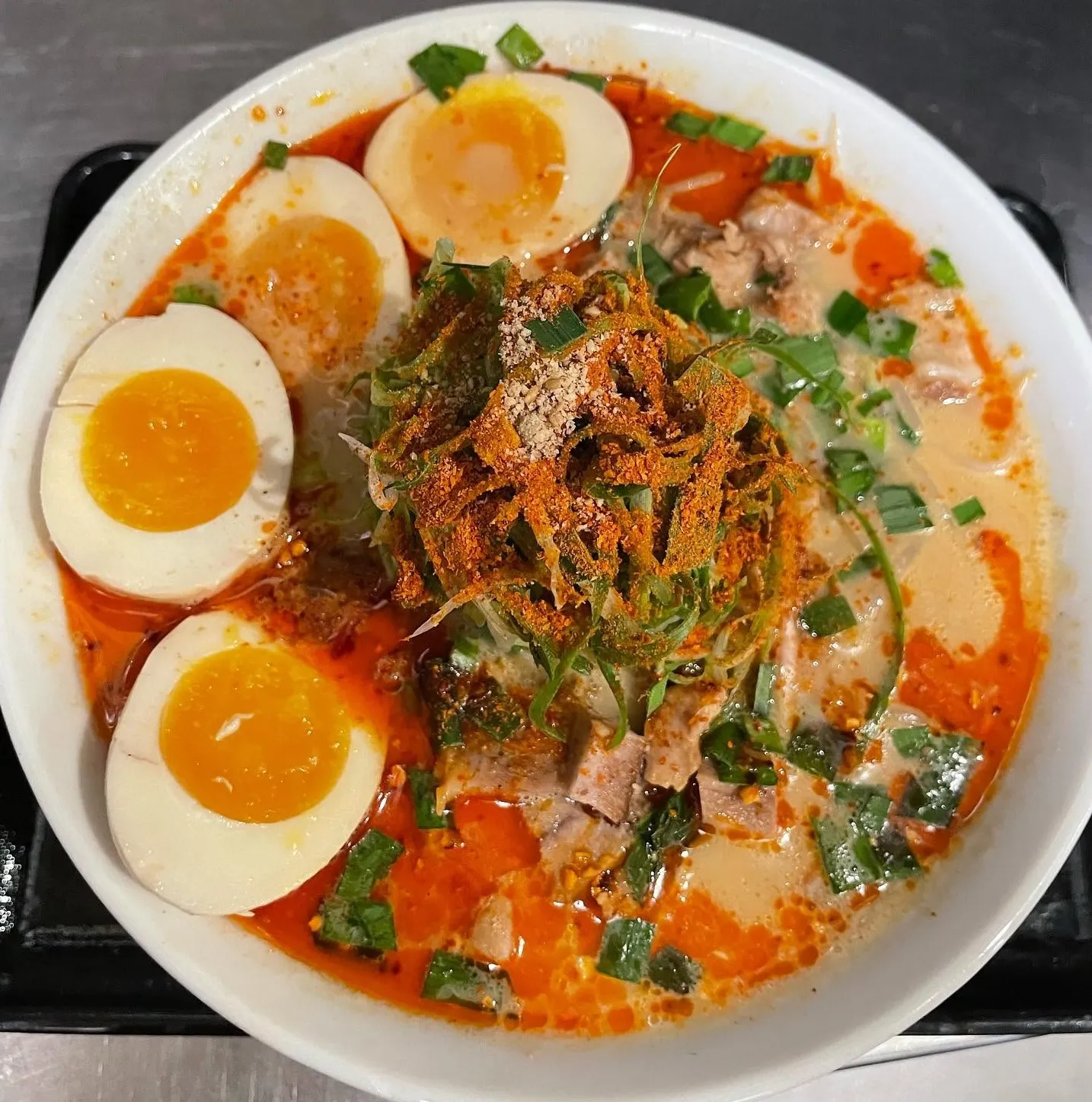 A bowl of spicy ramen noodles Costa Mesa topped with four halved soft-boiled eggs, sliced green onions, tender pork pieces, chili powder, and a creamy, orange-tinged broth with oil droplets.
