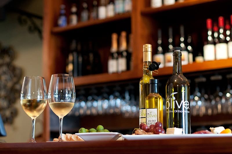 Two glasses of white wine, bottles of olive oil, and a plate with cheese, grapes, olives, and bread are set on a counter—perfect for anyone exploring things to do in Costa Mesa among vibrant shelves of wine bottles and glassware.