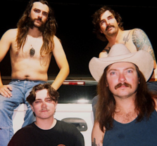 Four men with long hair and mustaches pose together; one is shirtless, another wears a tank top and cowboy hat, another has tattoos, and one wears a black shirt. They are standing by the back of a white pickup truck at night.