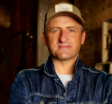 A man wearing a light-colored baseball cap and a denim jacket stands indoors, looking at the camera with a slight smile. The background is dimly lit with wooden and rustic elements.