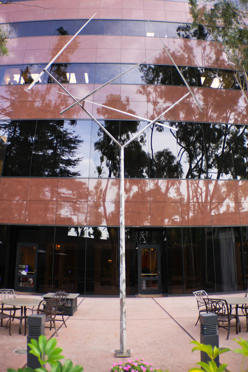 A tall metal sculpture with angular branches stands in front of a modern glass building, part of the Costa Mesa Art Walk, surrounded by patio tables, chairs, plants, and trees reflected in the building’s facade.