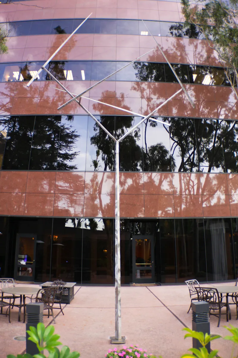 A tall metal sculpture with angular branches stands in front of a modern glass building, part of the Costa Mesa Art Walk, surrounded by patio tables, chairs, plants, and trees reflected in the building’s facade.