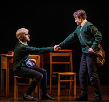 Two young men in green sweaters and school uniforms, one sitting and one standing, shake hands on stage with wooden desks and chairs behind them, suggesting a school setting and theatrical performance.