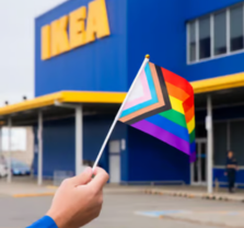 A hand holds a Progress Pride flag in front of an IKEA store with a blue and yellow exterior.