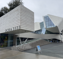 Modern, angular white building with large glass windows and unique geometric shapes. A sign near the entrance reads OCMA 3333. The sky is overcast and the sidewalk is wet.