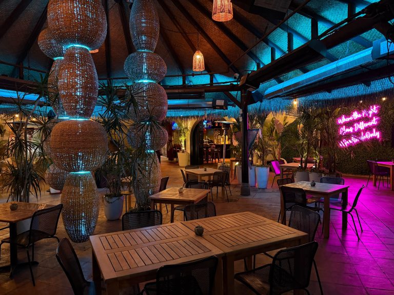 A cozy, tropical-themed restaurant with wooden tables, wicker chairs, hanging woven light fixtures, potted plants, and blue-purple mood lighting. A neon sign on the wall adds a vibrant touch to the relaxed atmosphere.