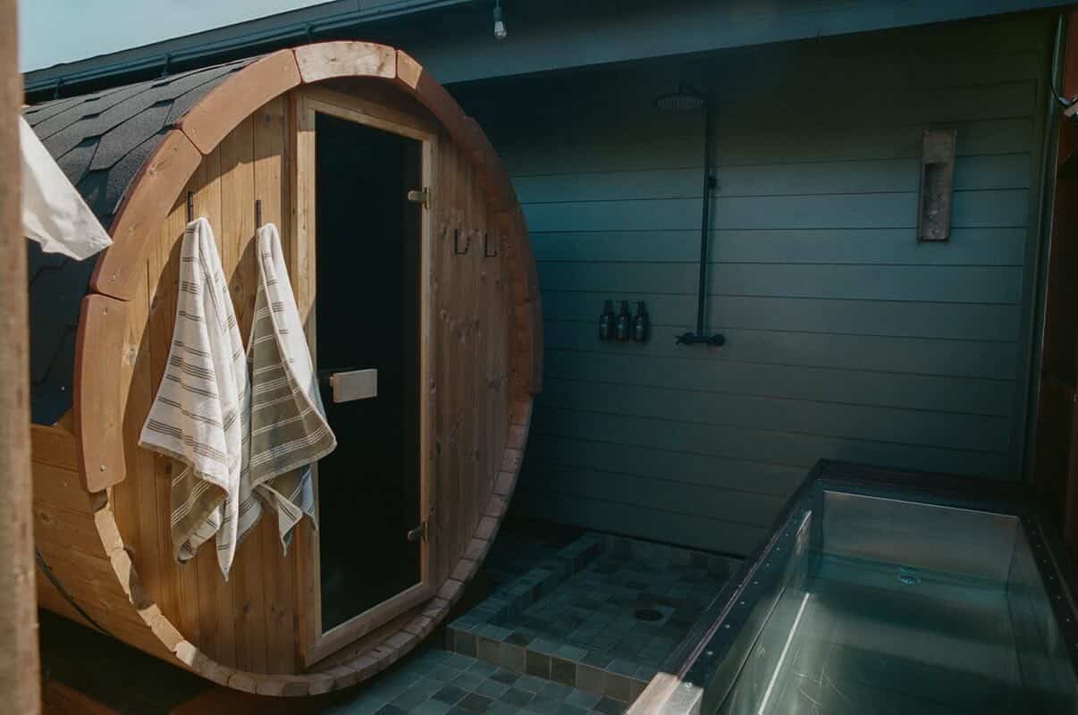 A wooden barrel sauna with two striped towels hanging on its door sits next to an outdoor shower and a rectangular soaking tub—one of the relaxing things to do in Costa Mesa—set against a paneled wall.