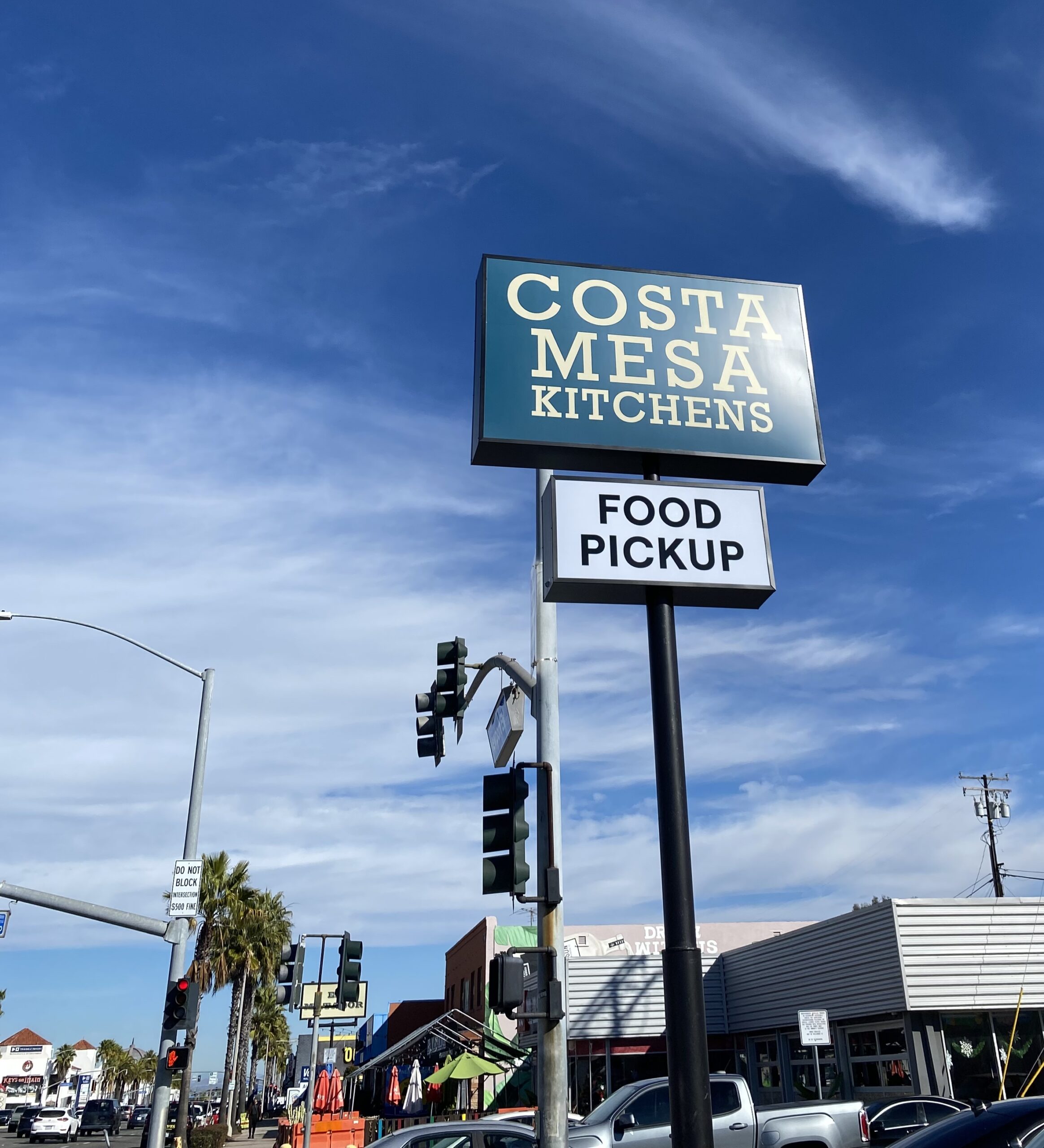 A street view shows a tall sign reading Costa Mesa Kitchens with a Food Pickup sign below it, set against a blue sky and surrounded by buildings, traffic lights, and palm trees.