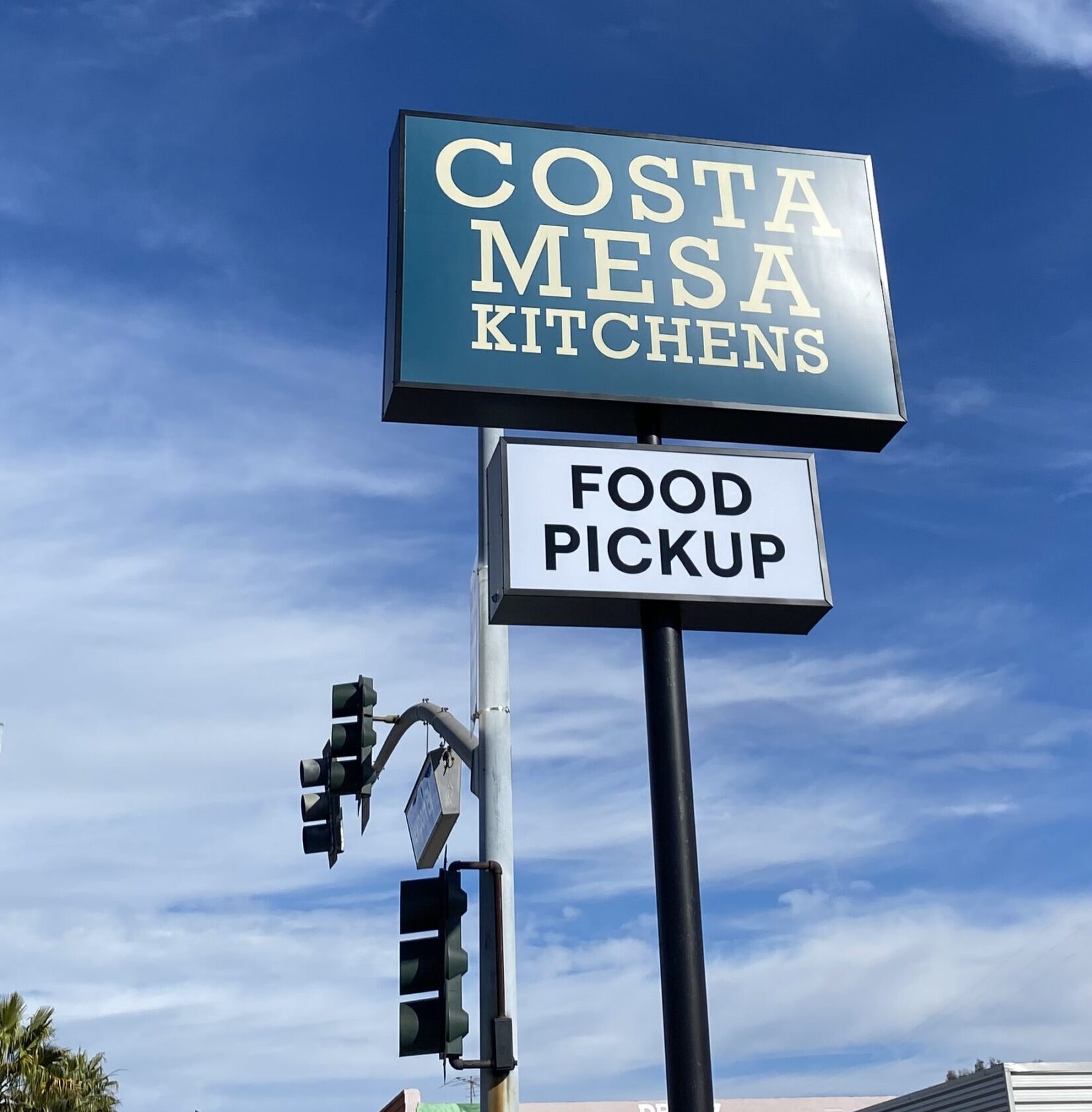 A street sign reads Costa Mesa Kitchens above a smaller sign that says Food Pickup, with traffic lights, blue sky, and clouds in the background.