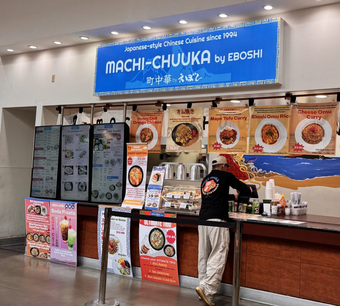 A man stands at the counter of Machi-Chuuka by Eboshi, a Japanese-style Chinese cuisine food stall with a blue sign, colorful menu displays, and food posters on the front wall.