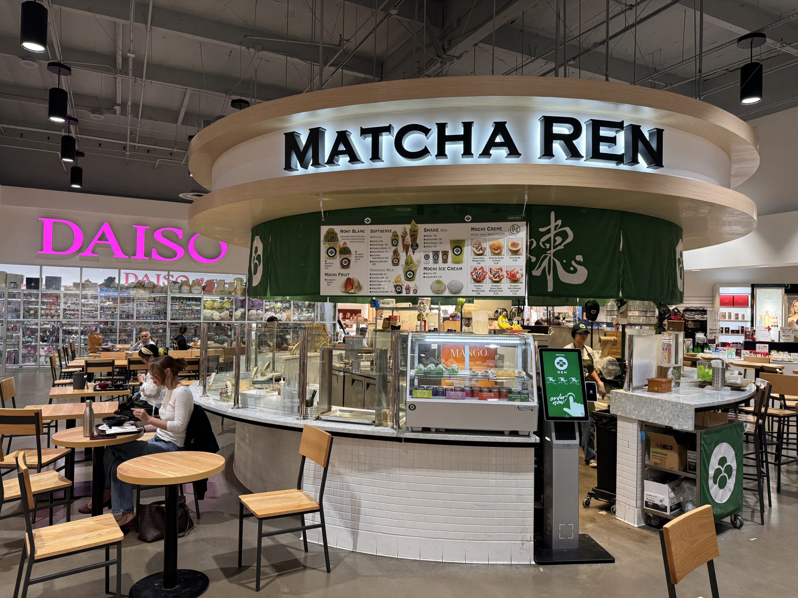 A modern matcha café called MATCHA REN with a menu above the counter, seating area with wooden chairs and tables, and a Daiso store visible in the background. A few people are seated and dining.