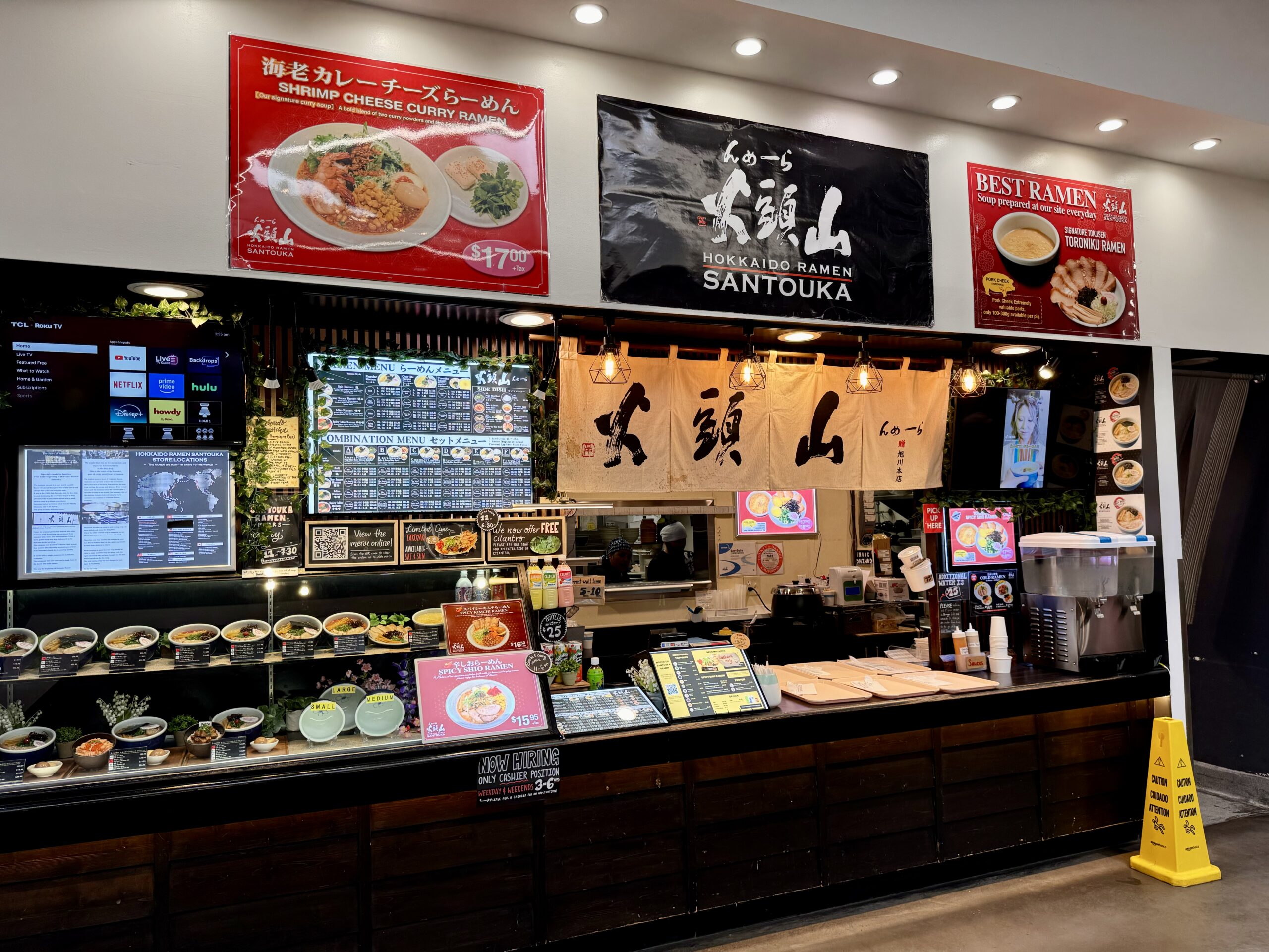A Japanese ramen restaurant counter with a menu display, food replicas, and large banners reading Hokkaido Ramen Santouka. Colorful menu boards show ramen dishes and prices above the counter.