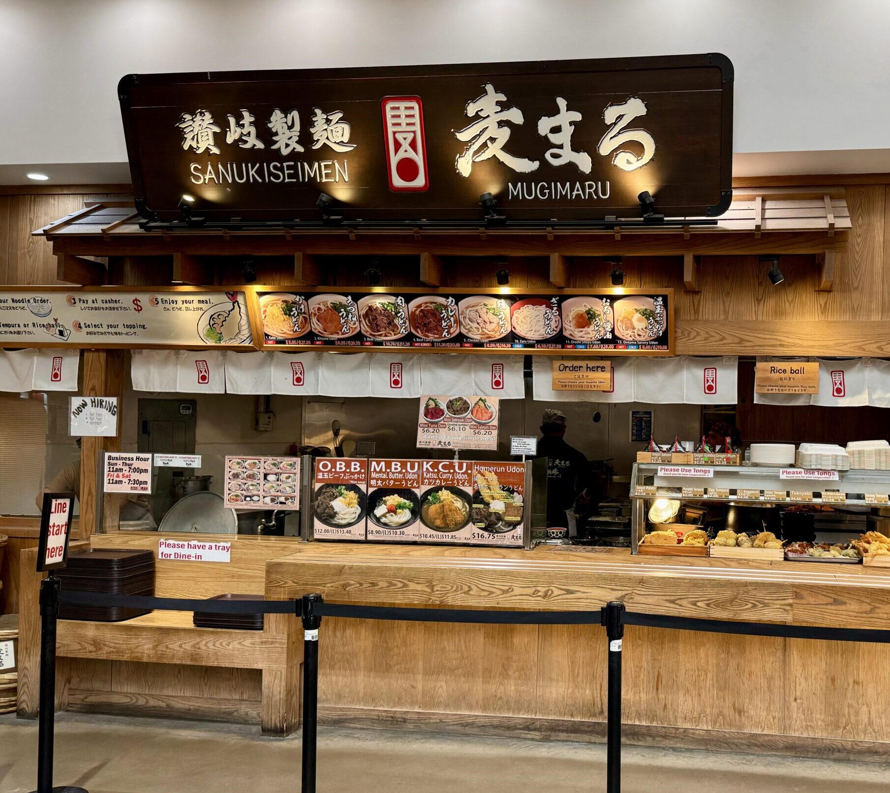 A Japanese noodle restaurant counter with a wooden facade, overhead menu boards displaying various udon dishes, and a sign reading Sanukiseimen Mugimaru. Food trays and utensils are arranged on the counter.