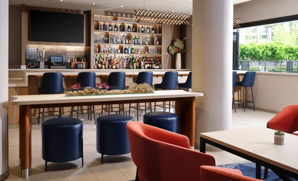 A modern hotel bar with a large liquor display, blue barstools, a TV above the bar, tables with red and blue seating, and large windows letting in natural light.