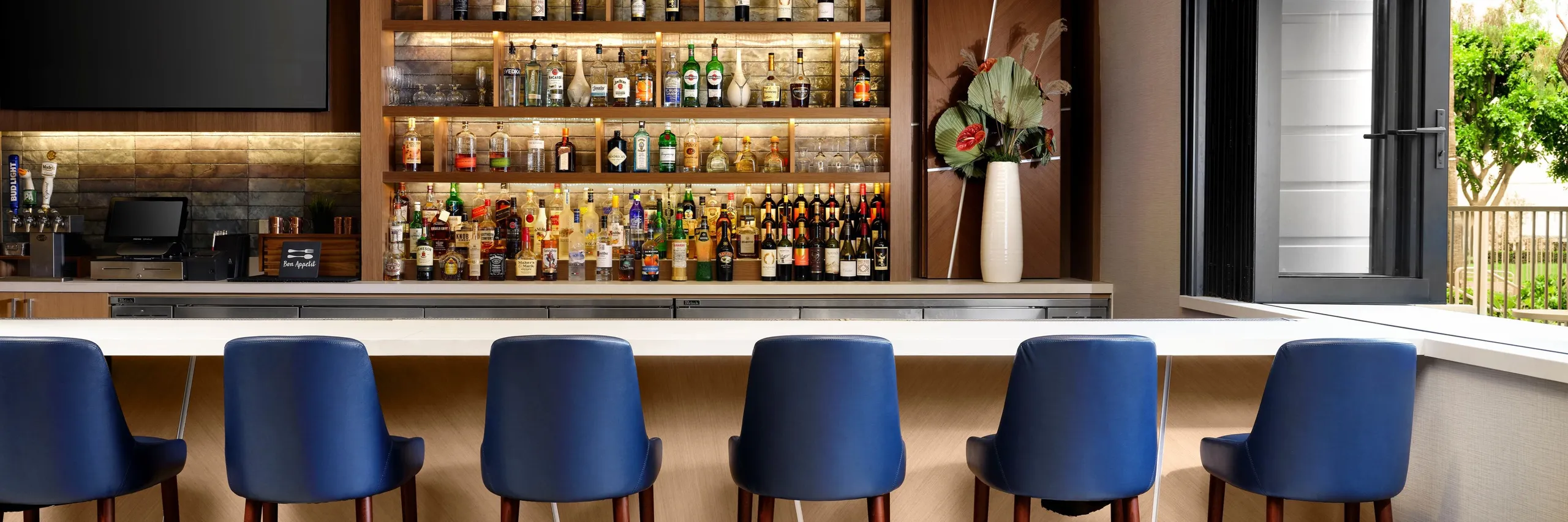 Modern bar with a row of six blue chairs at a white counter, a large display of various bottles on shelves behind, and a tall vase with flowers on the right side. A window on the right lets in natural light.