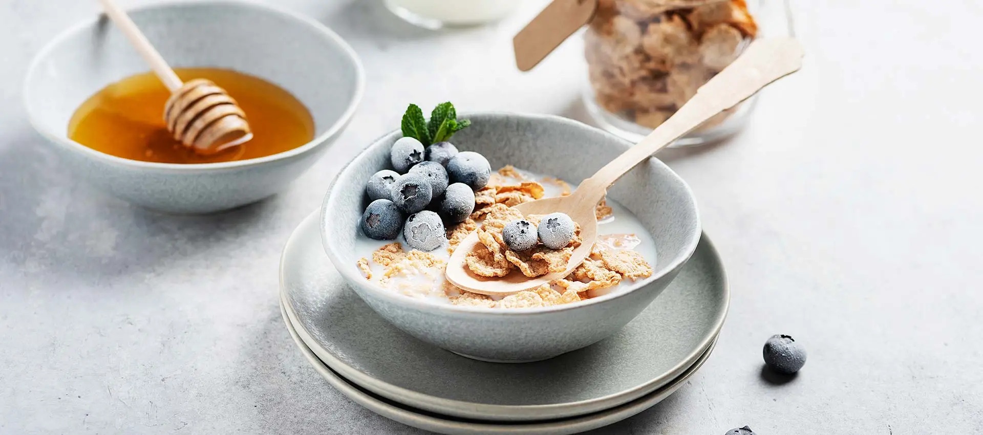 A bowl of cereal with milk and blueberries, garnished with a mint sprig, sits on stacked plates. A spoon rests in the bowl. Nearby are a bowl of honey with a dipper, a jar of cereal, and scattered blueberries.
