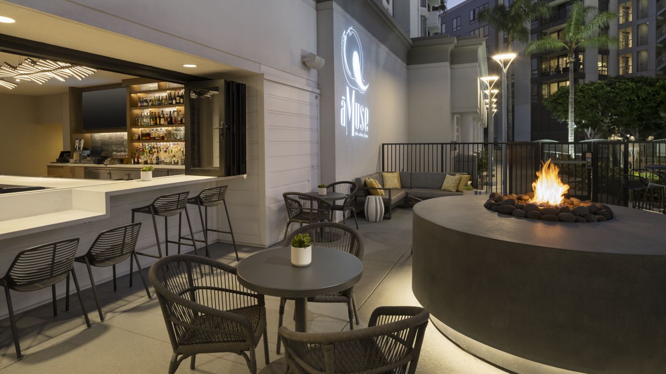 Modern outdoor lounge with a circular fire pit, black wicker chairs, small round tables, and a sleek bar area. Warm lighting and greenery create a cozy, inviting atmosphere in an urban setting.
