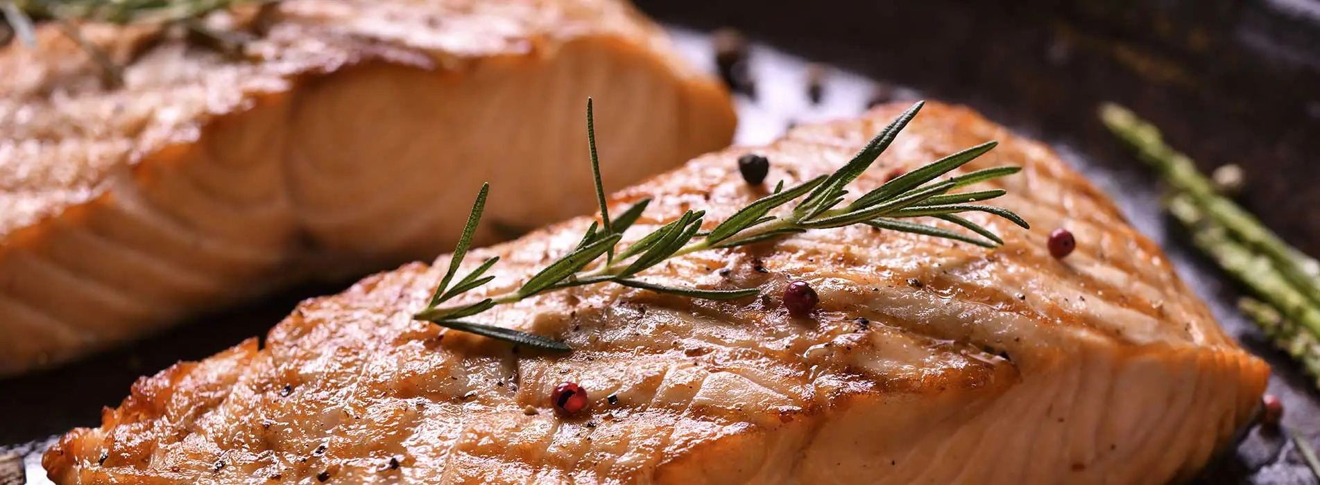 Close-up of a cooked salmon fillet topped with a sprig of fresh rosemary and peppercorns, with grill marks visible. Another piece of salmon and a green asparagus spear are blurred in the background.