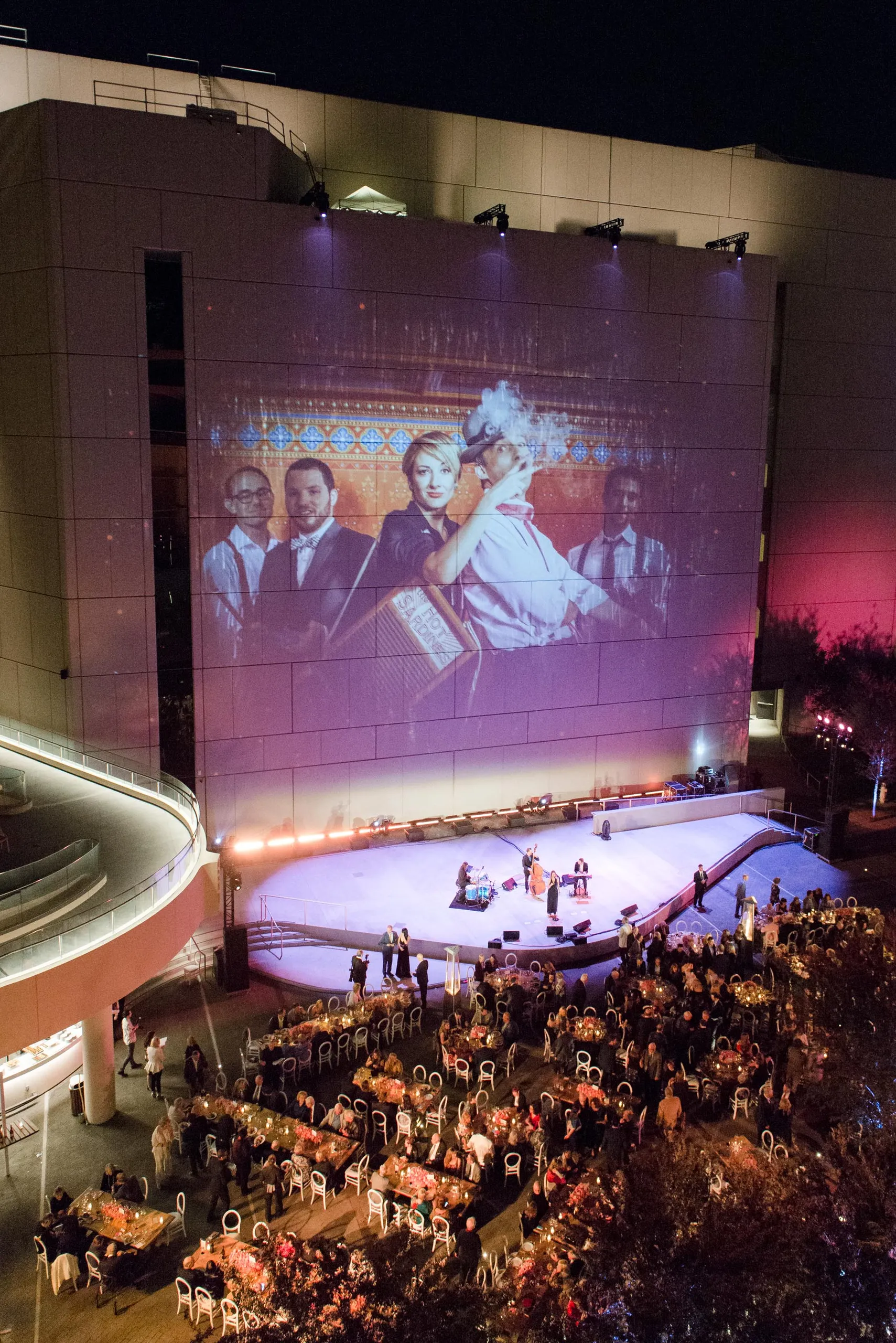 An outdoor event at night features guests seated at round tables facing a lit stage, set against a large building. As one of the premier meeting venues Costa Mesa offers, the scene includes a projected image of four people in vintage attire, two kissing.