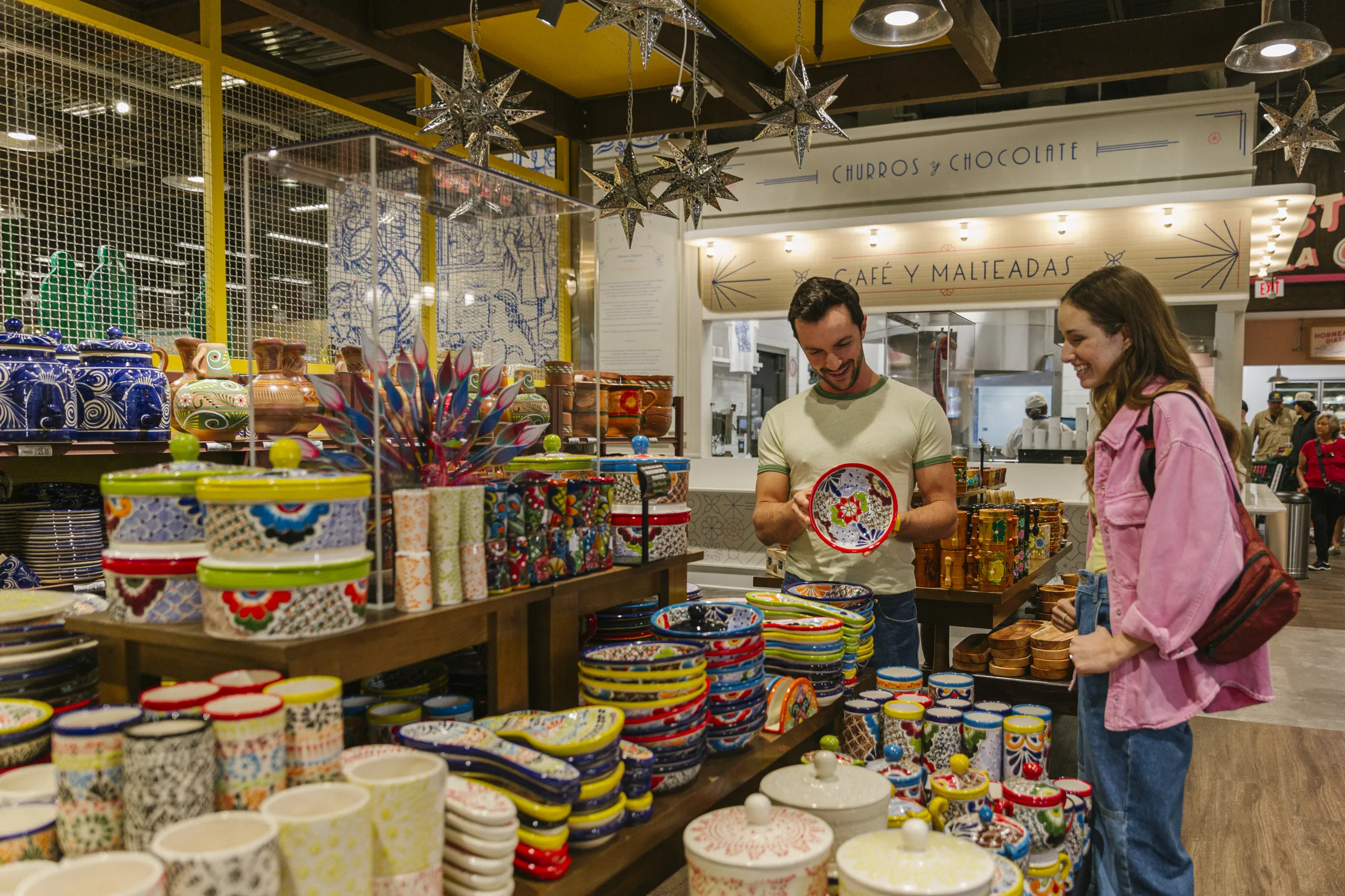 A man and woman smile while browsing colorful ceramic dishes at a market. Brightly painted bowls and plates are displayed around them, with star decorations hanging above.