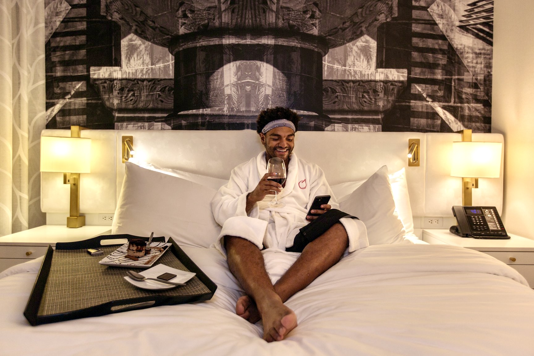 A person in a bathrobe sits on a hotel bed at one of the hotels Costa Mesa offers, smiling at their phone and holding a glass of wine. A tray with dessert and utensils rests on the bed, surrounded by warm lighting and modern decor.