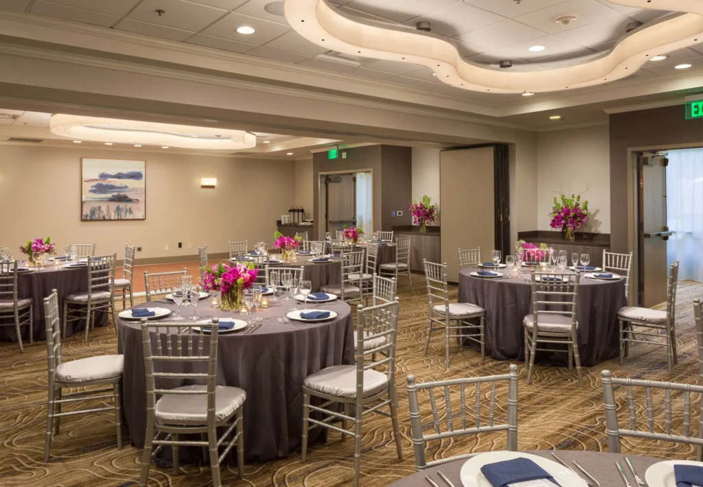 Elegant banquet room with round tables draped in gray linens, silver chairs, floral centerpieces, and place settings. Soft lighting and modern decor make it one of the most sophisticated meeting venues Costa Mesa has to offer.