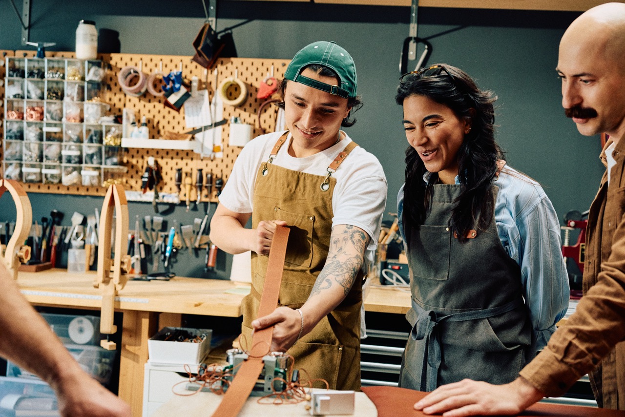 Three people in a workshop, wearing aprons, smile and examine a strip of leather. Tools and supplies hang on a pegboard in the background—a fun option if you're searching for creative things to do in Costa Mesa.