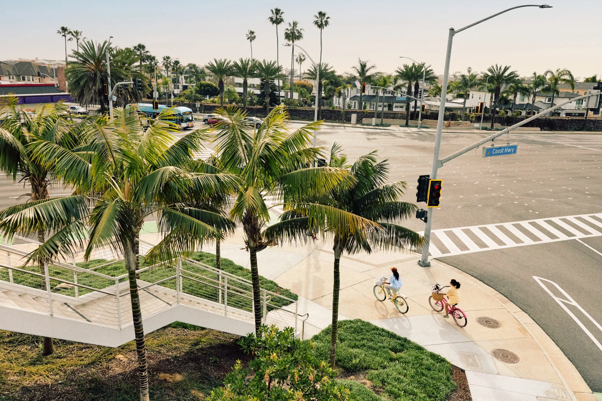 Two people ride bikes near palm trees at a sunny, empty intersection with crosswalks and a “Coast Hwy” street sign. Buildings and more palm trees line the background under a clear sky.