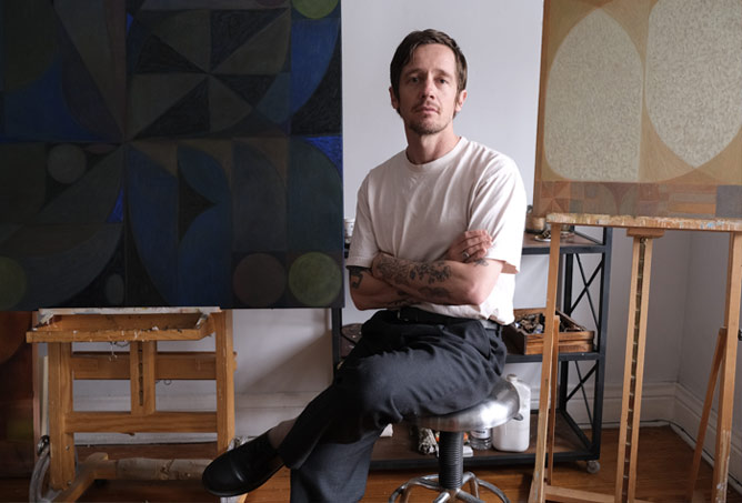 A man with tattoos, wearing a white t-shirt and dark pants, sits on a stool with arms crossed between two abstract paintings in an art studio.