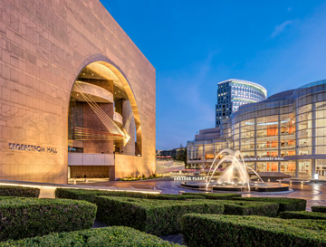 A modern performing arts center at dusk, featuring Segerstrom Hall with a large arched entrance, a glass-walled theater, a lit fountain, and manicured hedges in the foreground.