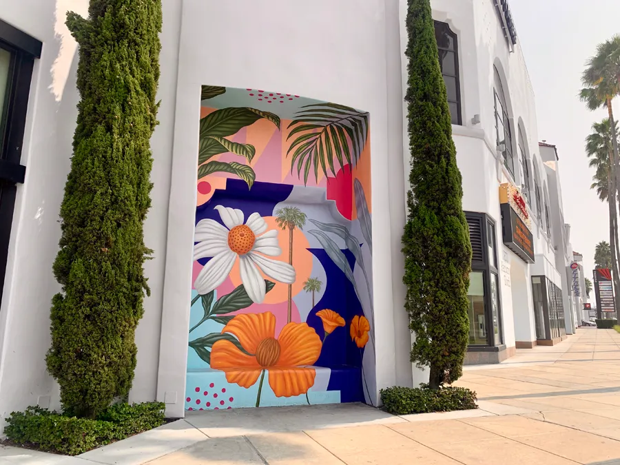 A colorful mural of large flowers and palm trees is painted on a recessed wall between two tall, slender cypress trees on a white building along a sunny, palm-lined sidewalk.