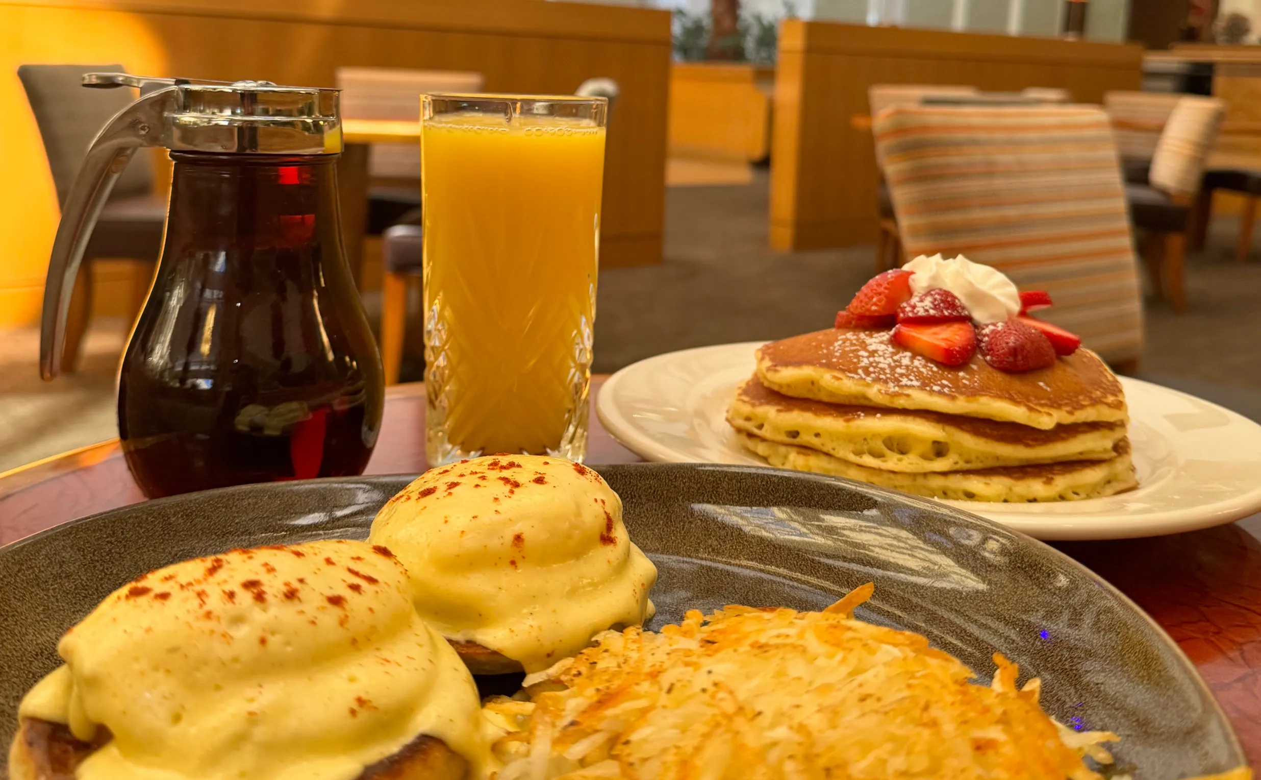 A breakfast table with eggs Benedict, hash browns, a stack of pancakes topped with strawberries and whipped cream, a glass of orange juice, and a syrup dispenser in a cozy restaurant setting.