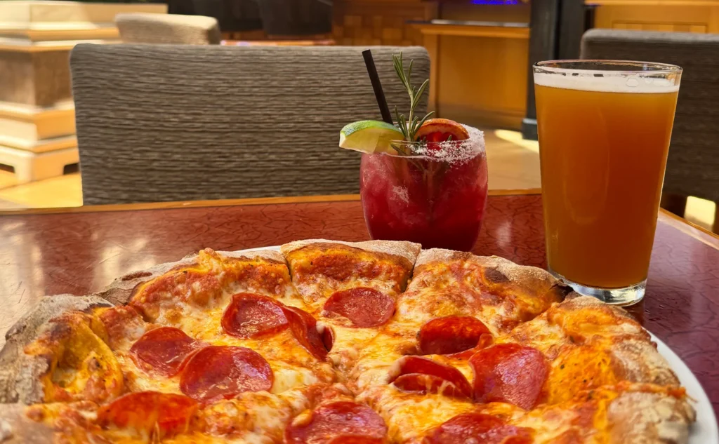 Pepperoni pizza, a ruby red cocktail with lime and rosemary, and a glass of amber beer on a table.