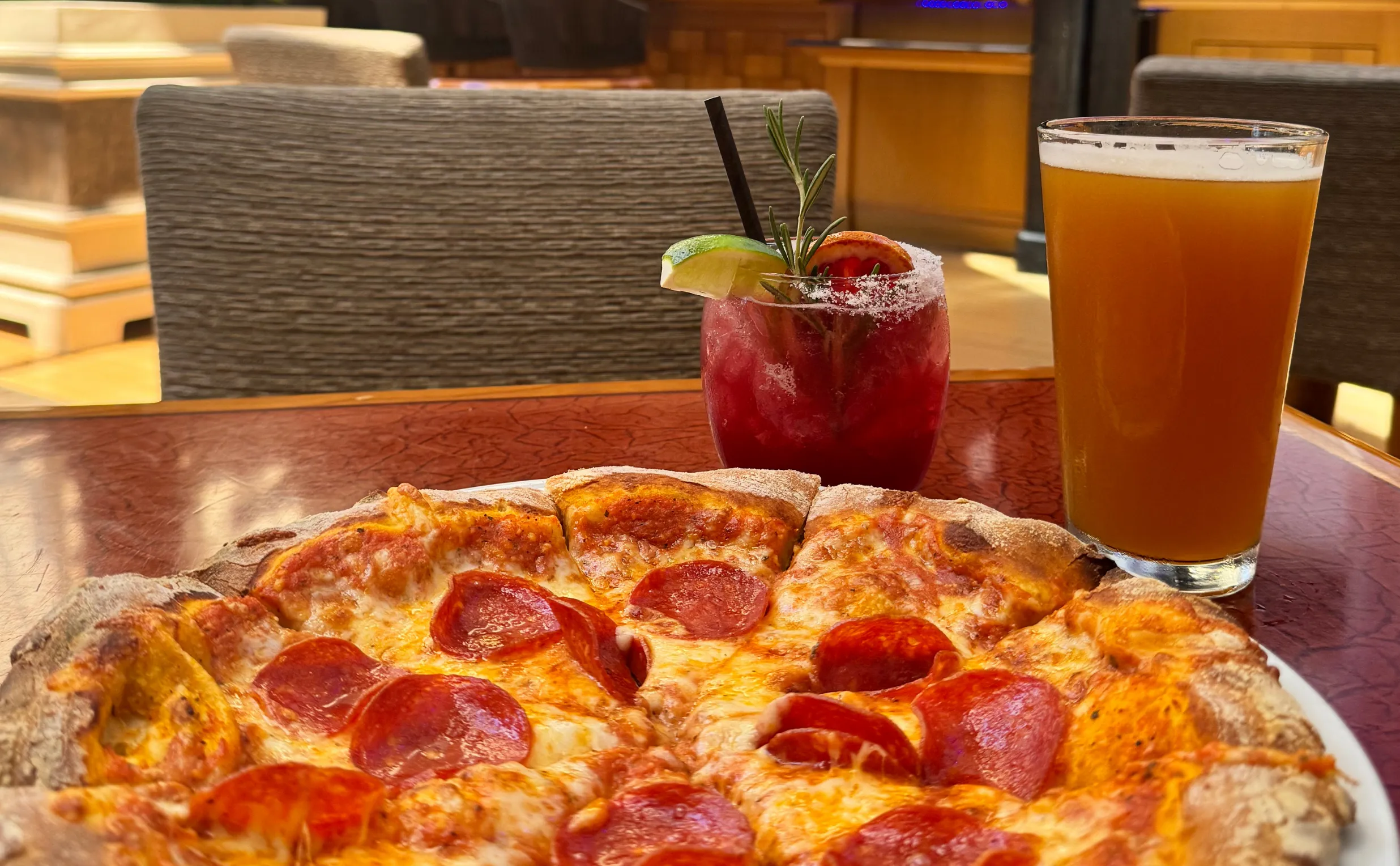 Pepperoni pizza, a ruby red cocktail with lime and rosemary, and a glass of amber beer on a table.