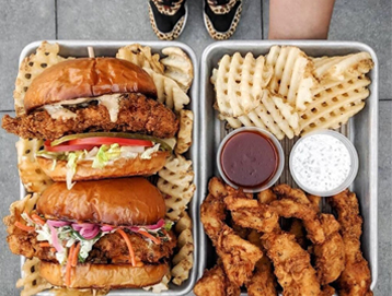 Two trays of food: one with two fried chicken sandwiches, lettuce, tomato, and waffle fries; the other with fried chicken tenders, waffle fries, two dipping sauces, and a persons feet in leopard print shoes visible at the top.