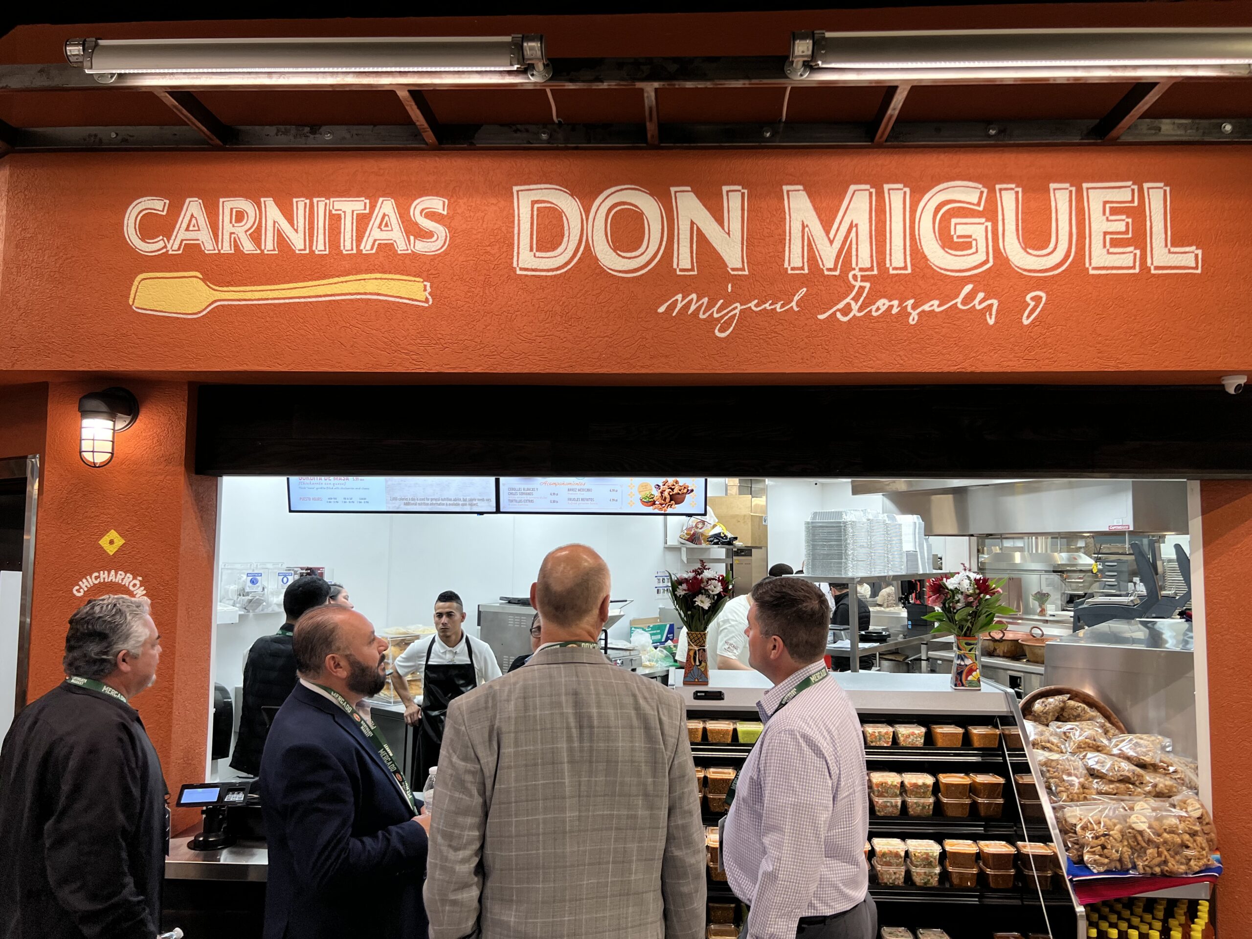 Four men stand in front of a counter at Carnitas Don Miguel, a food establishment with a visible kitchen and shelves of packaged baked goods. The restaurant sign is prominently displayed above the counter.