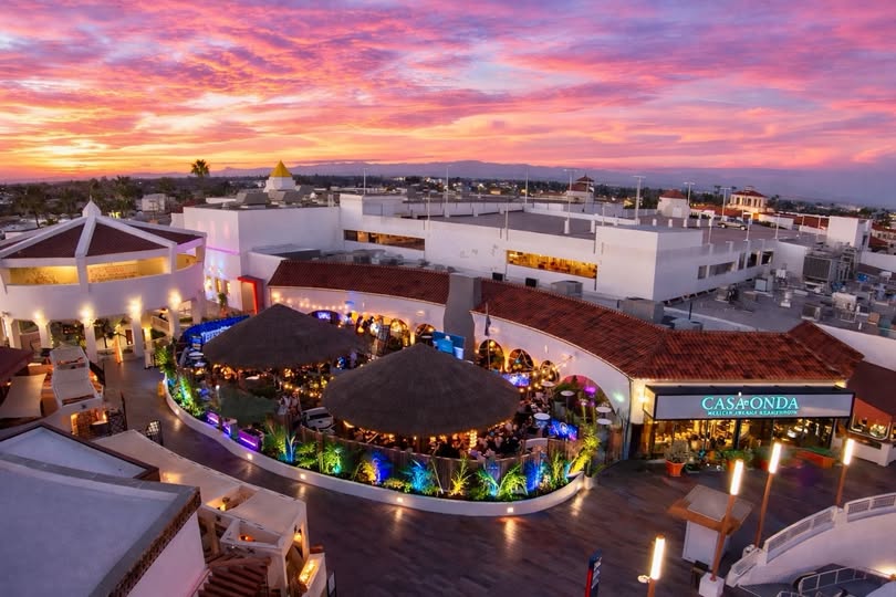 A vibrant outdoor shopping and dining area at sunset, featuring white buildings, a thatched-roof restaurant, colorful lighting, and a glowing pink and orange sky in the background.
