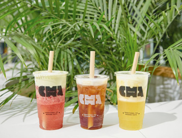 Three plastic cups of colorful drinks—red, brown, and yellow—each with a straw and the CHA logo, sit on a white surface with lush green plants in the background.