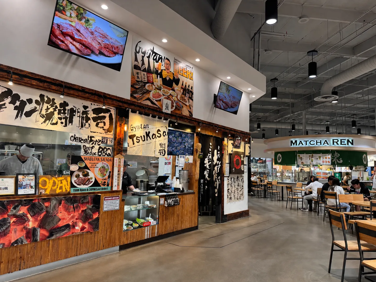 A Japanese food stall called Gyutan Tsukasa with colorful signs and menus stands inside a modern food court. Nearby, people are seated at tables near another eatery, Matcha Ren. The space is bright and spacious.