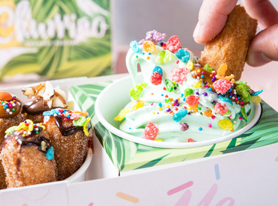A hand dips a churro into a cup of light green soft-serve ice cream topped with colorful sprinkles and cereal pieces. Nearby, more churros are served with chocolate sauce and sliced almonds.