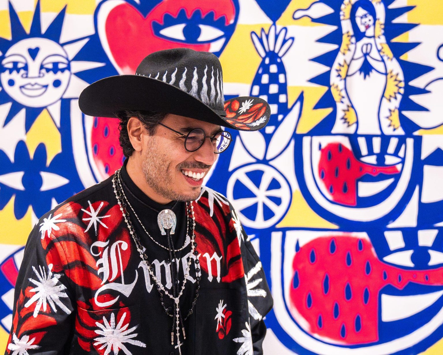 A smiling man in a black cowboy hat, glasses, and a colorful shirt stands in front of a vibrant, abstract mural featuring bold red, blue, yellow, and white patterns and stylized faces and religious imagery.
