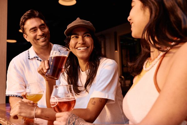 Three friends are smiling and talking at a bar, each holding a glass of beer. They appear to be enjoying each others company in a warm, casual setting.