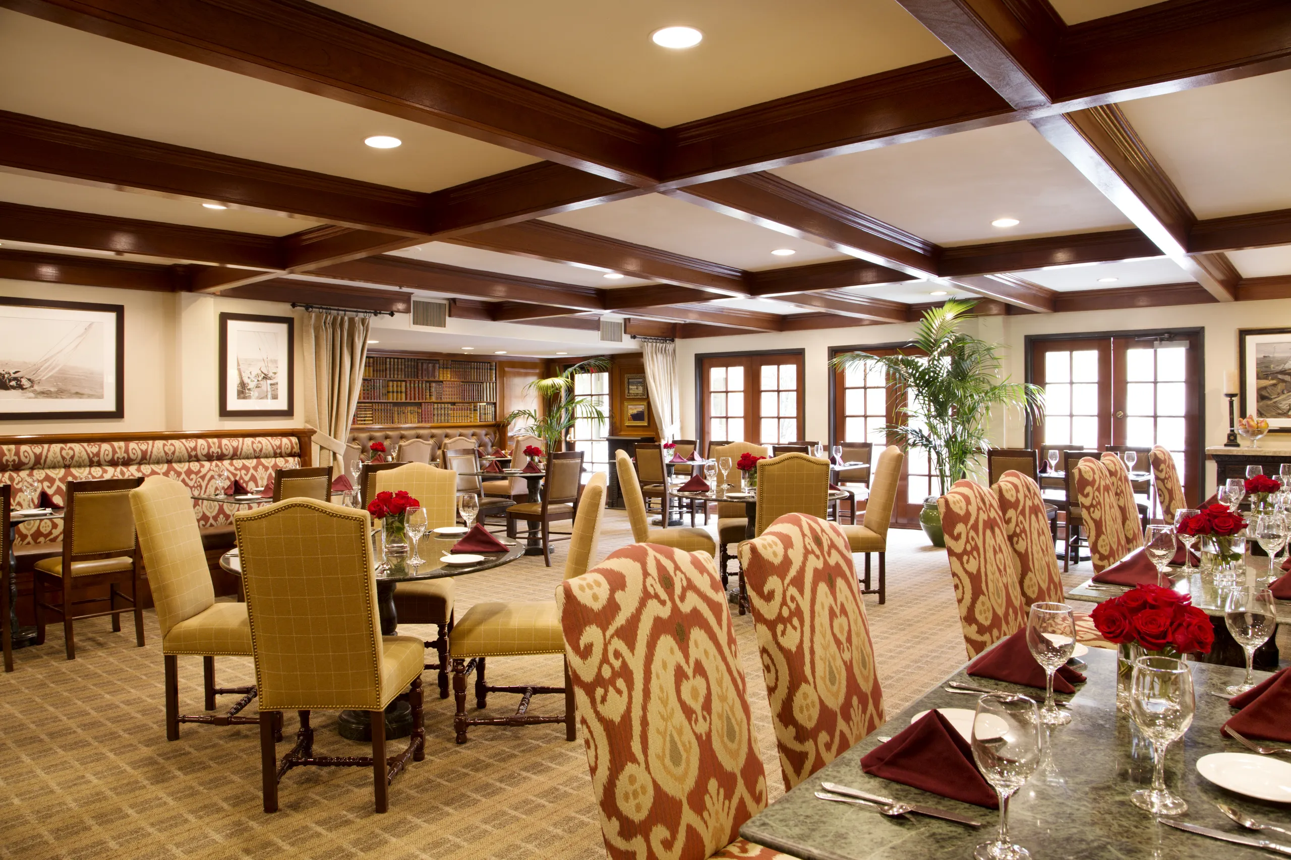 Elegant restaurant dining room with patterned chairs, set tables with red napkins and flowers, framed art on beige walls, bookshelves, large windows, and natural light. Warm, inviting atmosphere with classic decor.