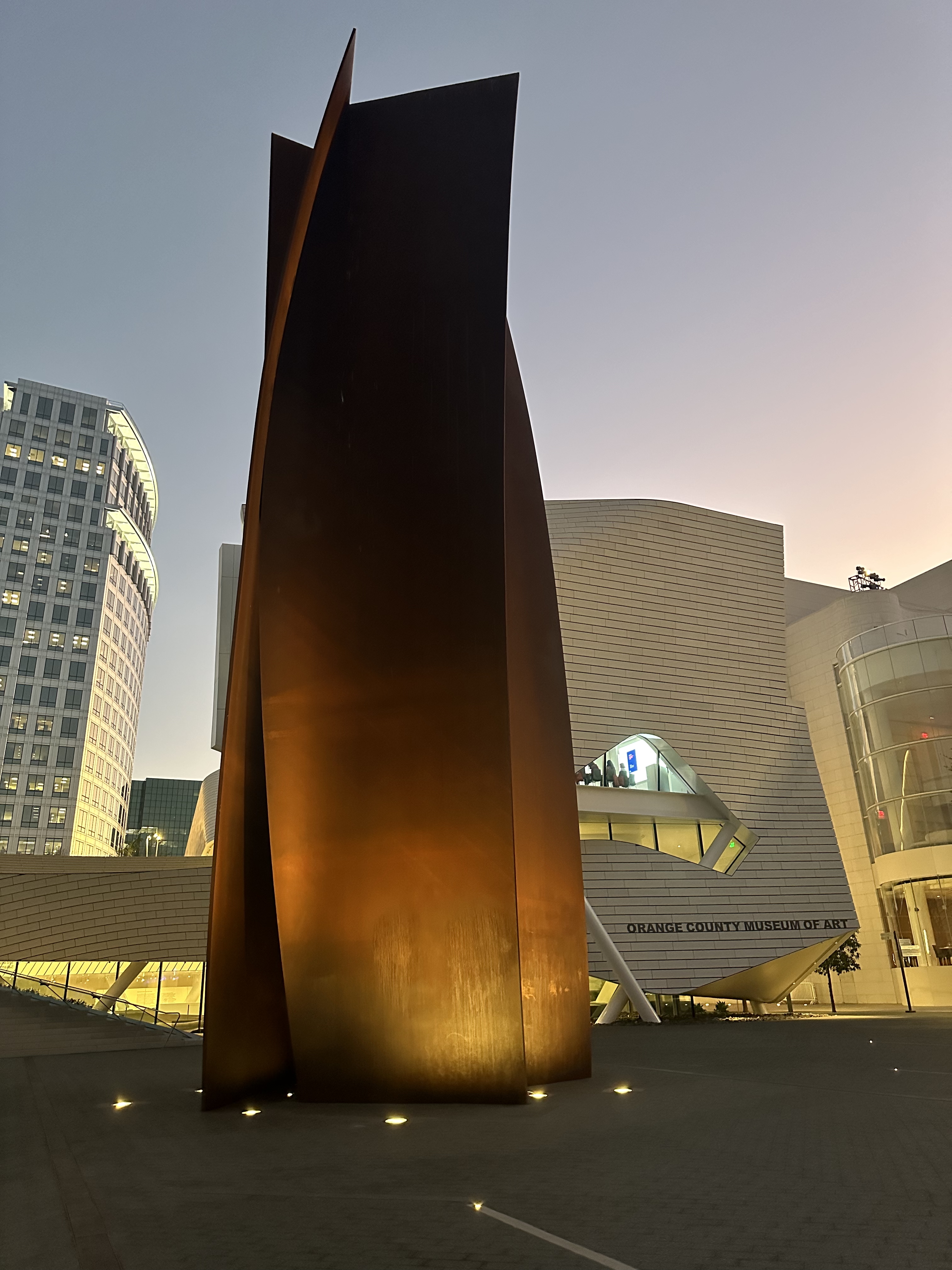 A tall, abstract rust-colored metal sculpture stands in front of the Orange County Museum of Art in Costa Mesa, City of the Arts, with modern glass and white buildings glowing softly at dusk.