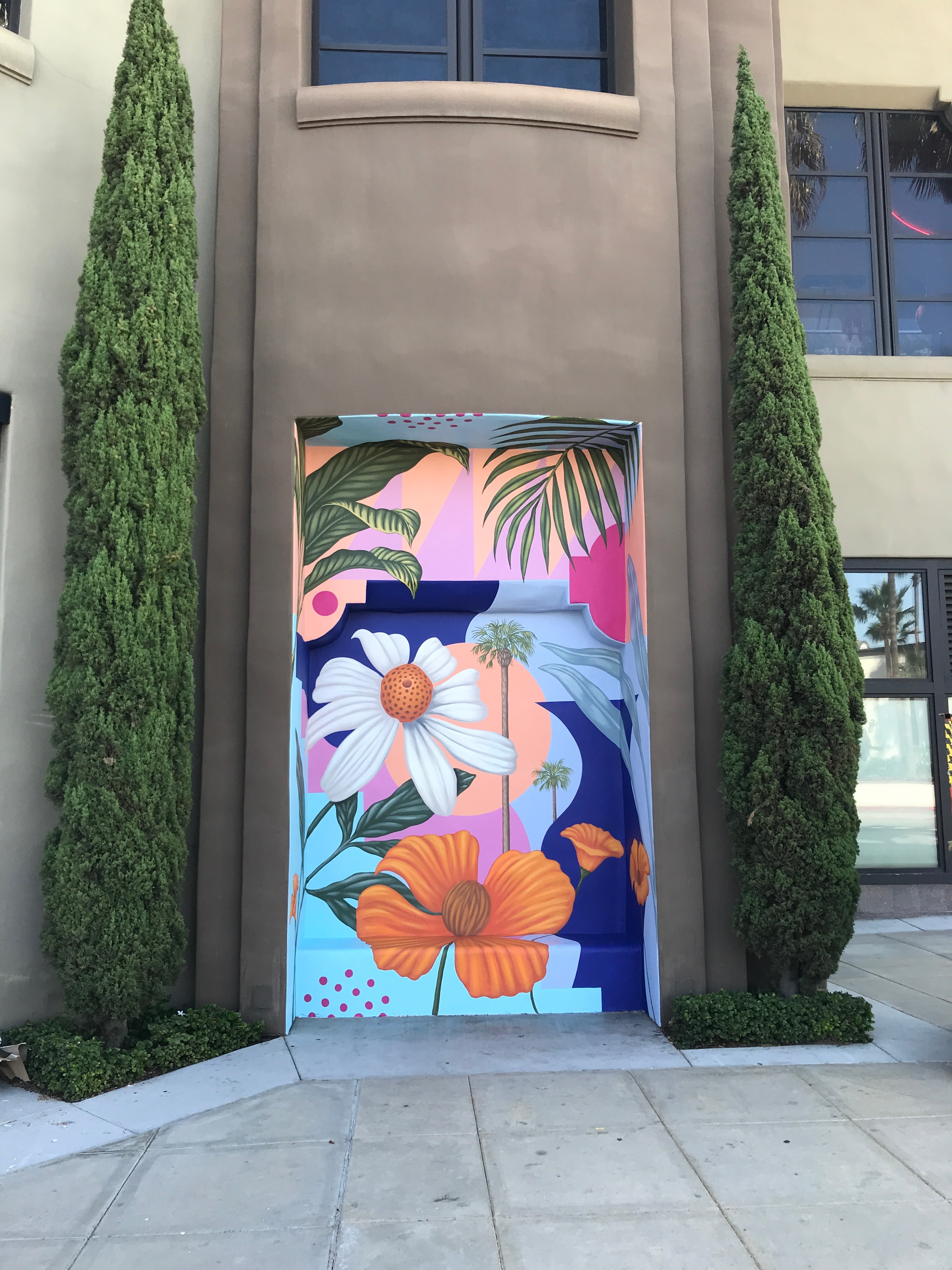 A colorful mural in Costa Mesa, City of the Arts, features large white and orange flowers with green leaves, decorating a recessed doorway between two tall, narrow cypress trees on the corner of a beige building.