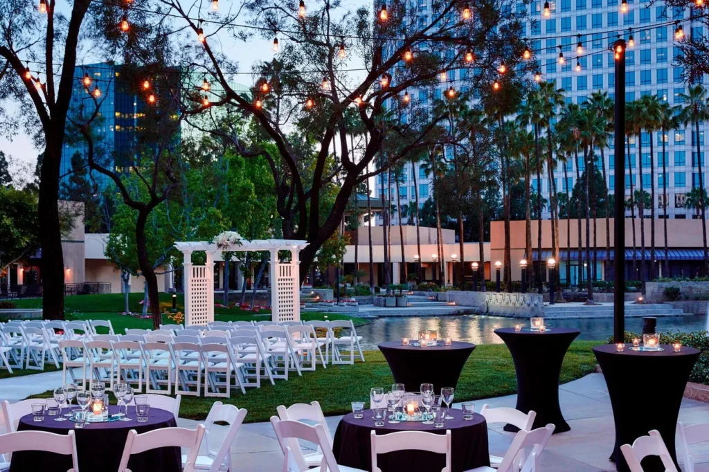 Outdoor event setting with round and rectangular tables, white chairs, string lights hanging from trees, and a small white pavilion—an inviting choice among event venues in Costa Mesa, surrounded by modern buildings and palm trees at dusk.