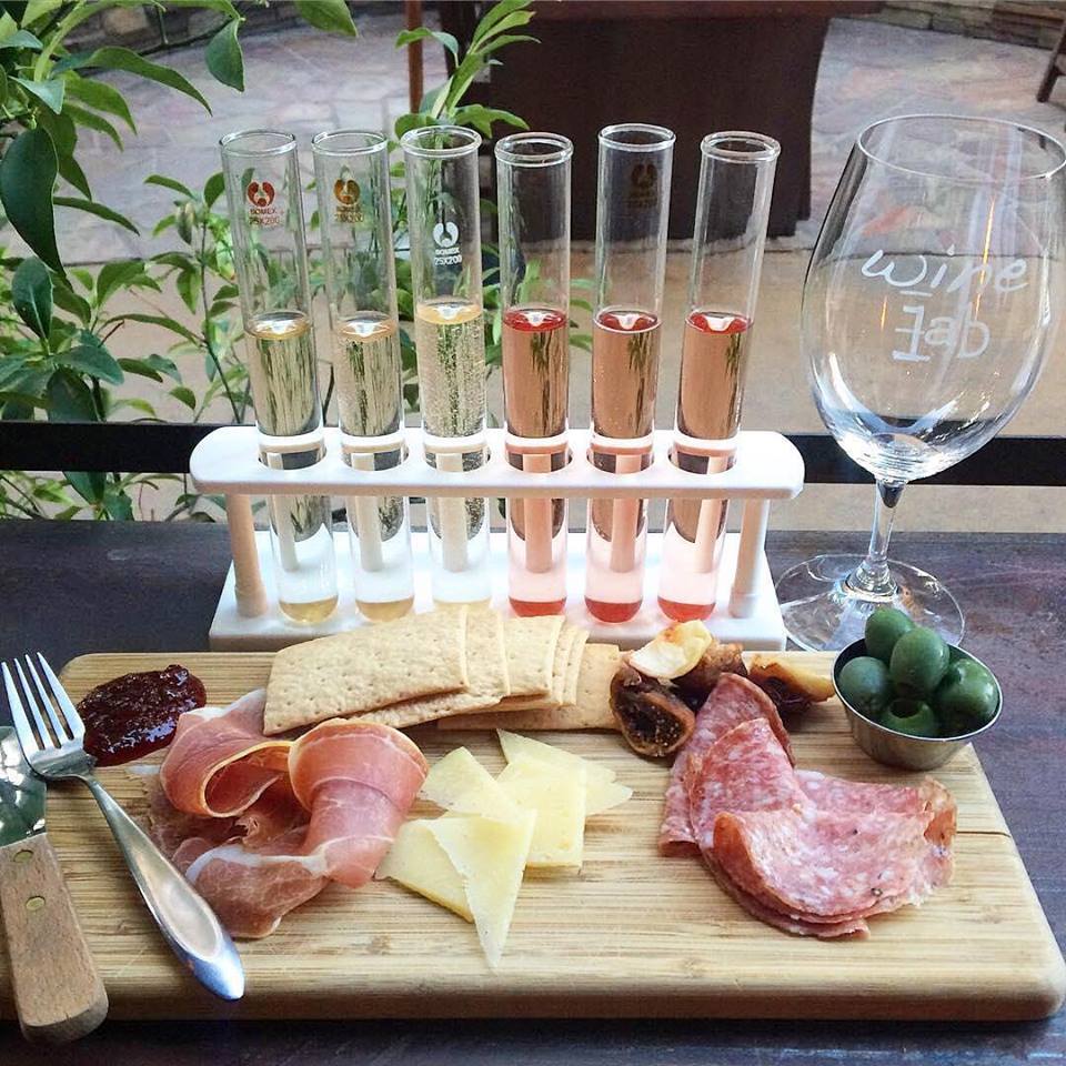 A charcuterie board with cured meats, cheese, crackers, figs, jam, and olives sits beside a wine glass and a rack of six wine-filled test tubes—one of the unique things to do in Costa Mesa—on a wooden table surrounded by greenery.