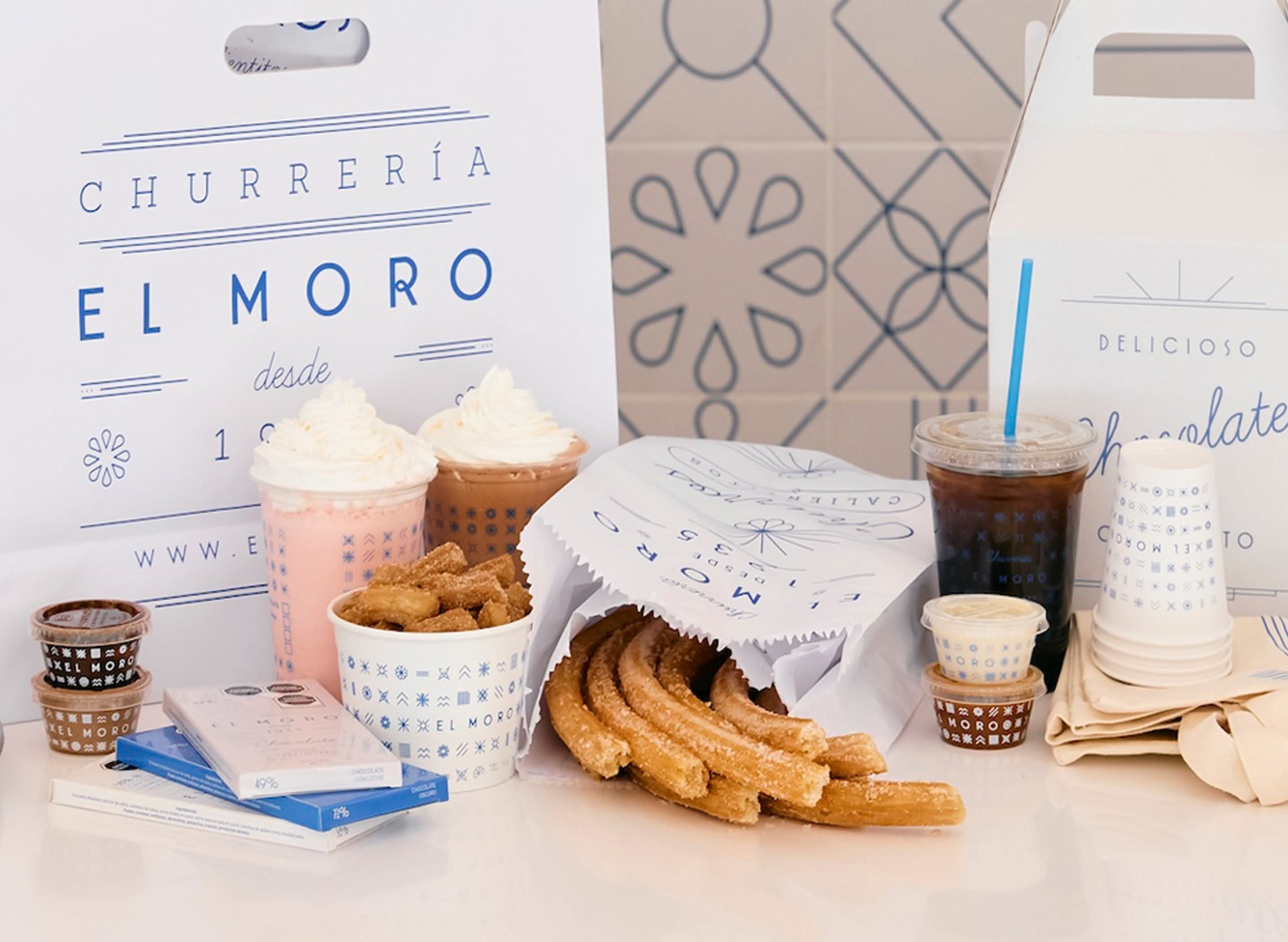 Assorted churros, drinks, and chocolates from El Moro are displayed on a white surface with branded packaging—a tempting lineup of some of the best desserts Costa Mesa has to offer, including hot chocolate, iced drinks, and dipping sauces.