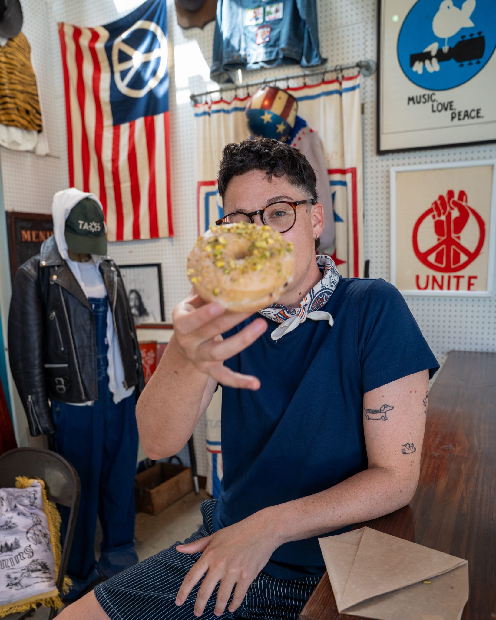 A person with short hair and glasses holds a pistachio-covered donut toward the camera, partially obscuring their face. They are seated in a vintage shop decorated with Americana and peace-themed posters and flags.